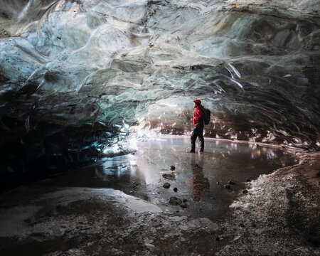 A man standing in an ice cave lit up with light, with clear contoured ice on the ceiling, walls of ice and frozen water ripples and waves.
