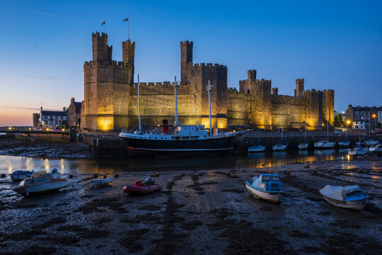 The historic Norman motte and bailey Caernarfon Castle, walls lit up at dusk, the estuary of the Afon Seiont river, boats moored.
