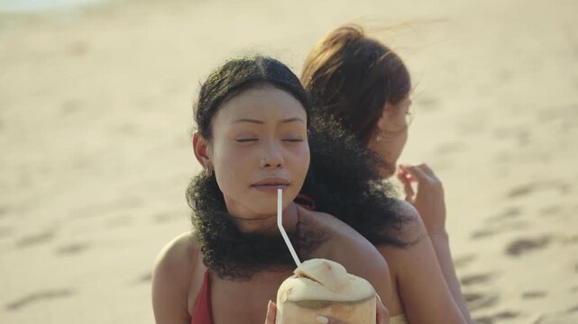 Two smiling multiethnic young women in swimwear enjoying fresh coconut drinks while relaxing on a sunny tropical beach during their summer vacation travel at sunset background.