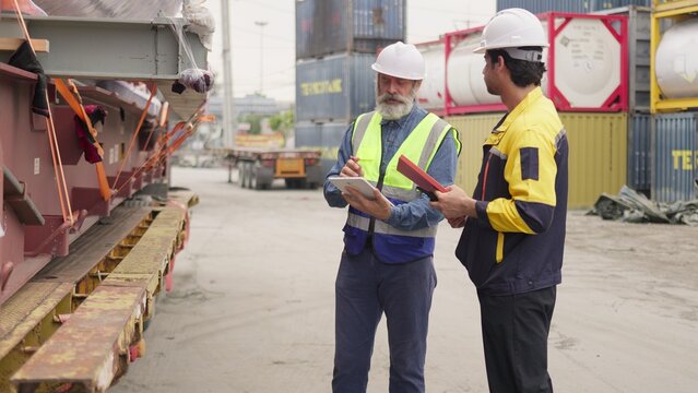 Two logistics workers inspecting secured cargo on a flatbed truck at a shipping terminal, collaborating on supply chain management using a digital tablet.
