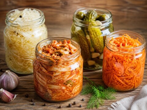 Assorted fermented foods in clear glass jars, including sauerkraut, kimchi, pickled cucumbers and fermented carrots, arranged on a rustic wooden table. Natural soft light and clean editorial food styl
