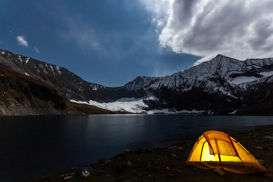 View of a glowing yellow tent on the shore of Ratti Gali Lake surrounded by snow-capped mountains under a starry night sky in Neelum, Azad Kashmir, Pakistan.
