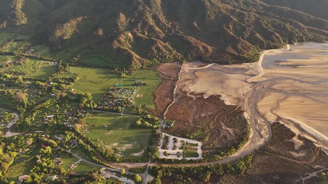 Sunrise over Marahau coastal village in Abel Tasman region South Island New Zealand with sandy estuary green fields and surrounding mountains glowing in warm morning light scenic landscape. Aerial