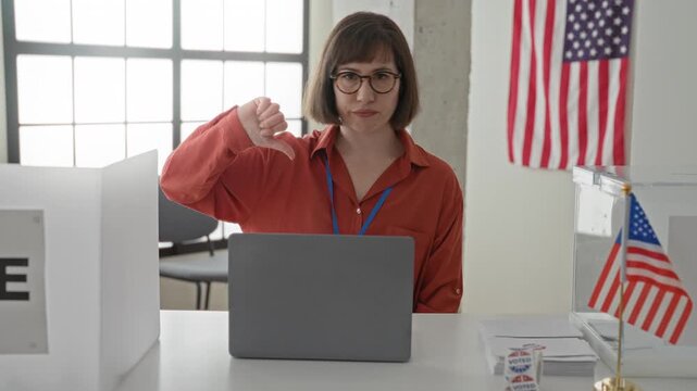 Woman with short hair and glasses at polling desk using laptop giving thumbs down gesture in voting building with ballot box and american flag visible; disapproval civic concern.