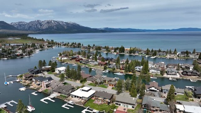 Luxury waterfront homes and boat docks in Tahoe Keys with calm canals leading to Lake Tahoe, surrounded by pine trees and snow capped Sierra Nevada mountains in California,USA. Aerial backwards.