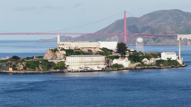 Alcatraz Island in San Francisco Bay, California, USA, with historic prison buildings, lighthouse and water tower, set against Golden Gate Bridge and coastal hills in calm scenic light. Aerial drone v