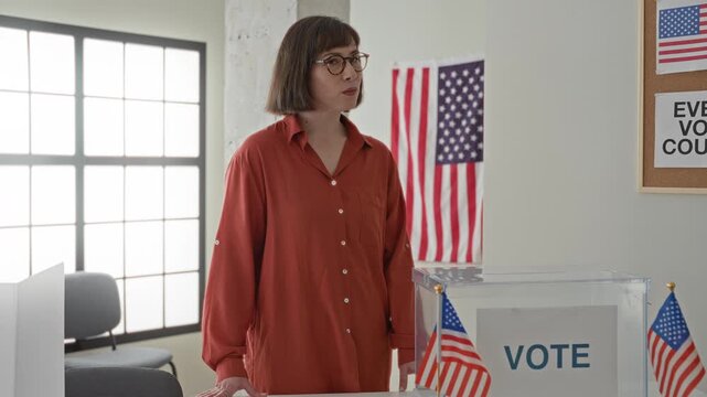 Young hispanic woman wearing red shirt placing hand on transparent ballot box in voting building with us flags and vote sign; civic duty hopeful.