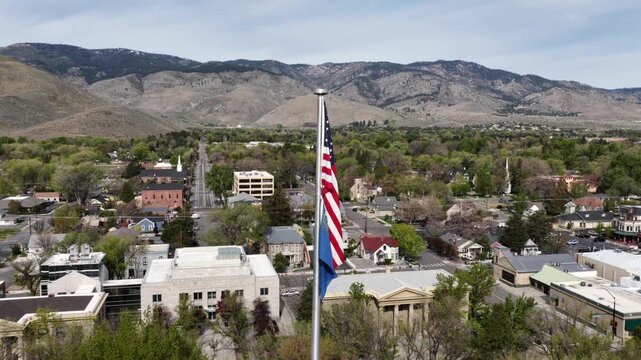 American and Nevada state flags waving above Carson City in residential neighborhood with tree-lined streets, civic buildings and mountain backdrop under clear daylight sky. Aerial orbit shot.
