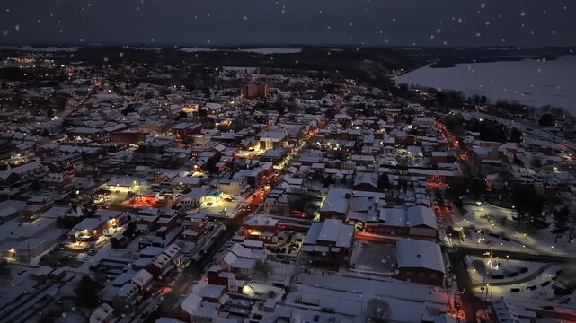 Aerial drone rise over snow-covered small town main street in United States at dusk, with warm streetlights illuminating traffic, row houses and quiet winter atmosphere during holiday season.
