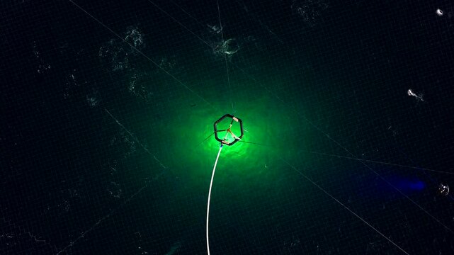 Glowing green underwater light in circular salmon farm pen during polar night in Norway. Aerial top-down view