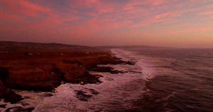 Rocky coastline and pacific ocean waves at Punta de Lobos during vibrant pink sunset in Pichilemu, Chile. Aerial forward, coipy space