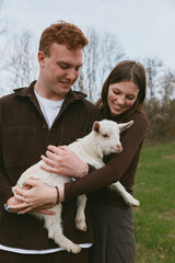 Fototapeta premium Joyful couple cradle a white goat kid in a sunlit field, showcasing tender bonding and rustic charm during a casual countryside moment, with gentle smiles and shared joy under blue sky