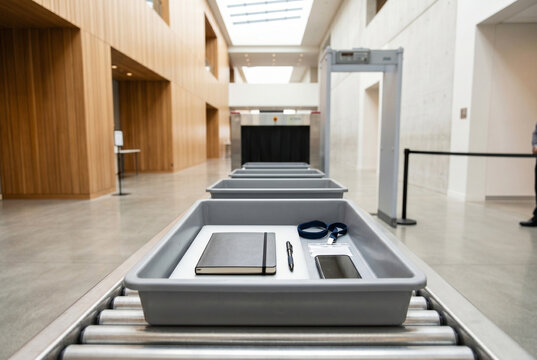 A security check point is set up inside a modern building. There are several trays on a conveyor belt with a notebook, pen, and a phone. The area is open and well lit.