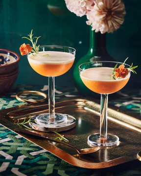 Two Elegant Floral Cocktails in Coupe Glasses with Rosemary and Marigold Garnish on Gold Tray