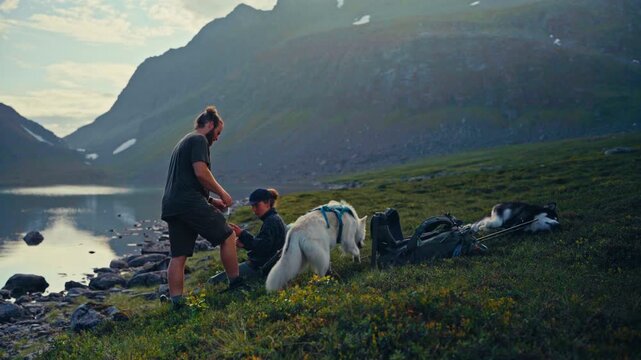 S&oslash;r Tverrfjord, Loppa, Norway - Two Hikers Rest by a Serene Lakeside as One Sprays Insect Repellent while Dogs Sit Beside them in a Scenic Mountain Landscape - Static Shot