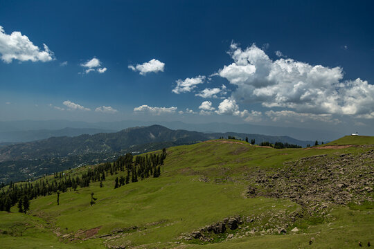 View of Ganga Choti peak featuring lush green meadows, rocky slopes and pine trees under a blue sky with white clouds in Bagh, Azad Kashmir, Pakistan.