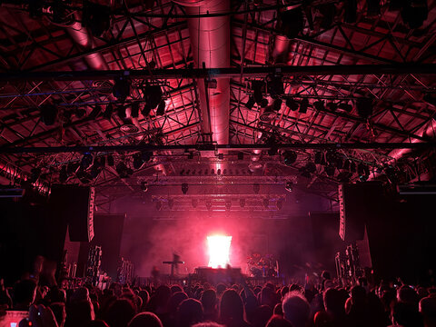 High-energy rock concert crowd gathered in a large, packed music venue. A band performs on stage under dramatic, colorful lighting, with hundreds of people cheering and raising their hands