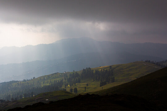 View of the majestic Ganga Choti peak with sunbeams breaking through dark storm clouds over lush green hills and pine trees in Bagh, Azad Kashmir, Pakistan.