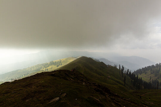 View of Ganga Choti mountain ridge with people standing on the green slopes under a heavy overcast sky in Bagh, Azad Kashmir, Pakistan.