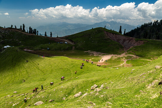 View of Ganga Choti peak with people on horseback traveling along a mountain trail through lush green meadows under a cloudy blue sky in Bagh, Azad Kashmir, Pakistan.