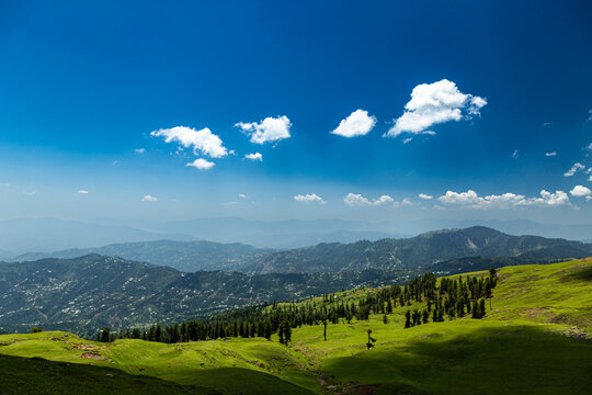 View of Ganga Choti peak with lush green meadows and scattered pine trees under a bright blue sky with white clouds in Bagh, Azad Kashmir, Pakistan.