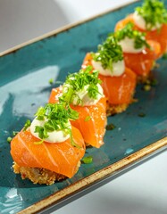 A close-up shot shows a row of salmon sushi bites topped with white sauce and green herbs on a teal rectangular plate. The sushi pieces are arranged in a diagonal line on the plate.