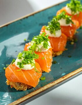 A close-up shot shows a row of salmon sushi bites topped with white sauce and green herbs on a teal rectangular plate. The sushi pieces are arranged in a diagonal line on the plate.