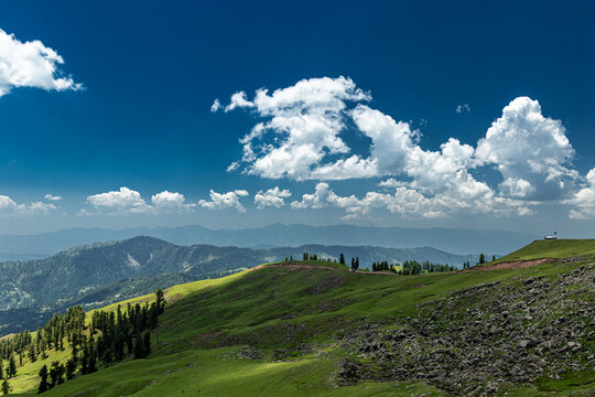 View of Ganga Choti peak and lush green meadows under a bright blue sky with fluffy clouds in Bagh, Azad Kashmir, Pakistan.