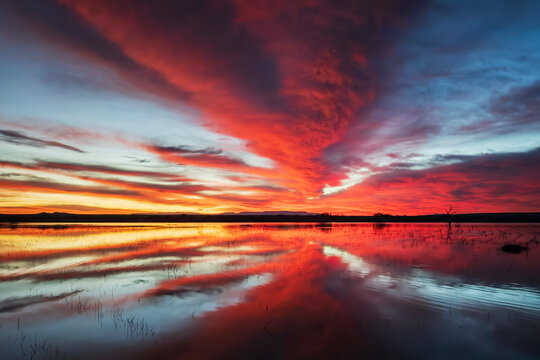 Orange and yellow glow on the horizon at sunset, reflected in a flat calm water surface, a dramatic sky.