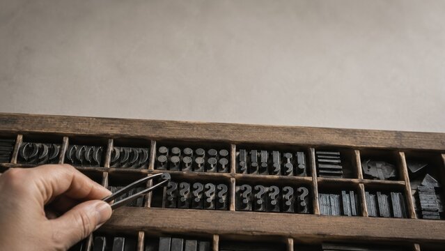 Hand using tweezers to select a metal question mark type block from inside a wooden letterpress drawer filled with vintage punctuation pieces