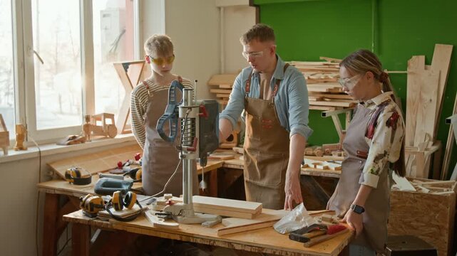 Young professional carpenter showing two adolescent kids how to use drilling press machine during woodworking lesson