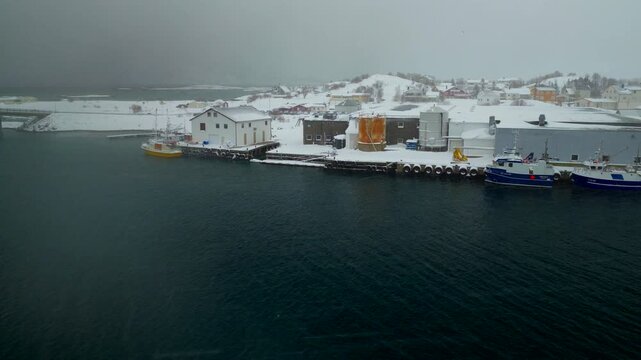 Snowy white Sommaroya with fishing boats moored in harbor, aerial as snow falls