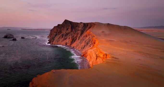 Drone flying over stunning red cliffs and desert landscape of Paracas National Reserve lit by the setting sun in Peru. Aerial forward