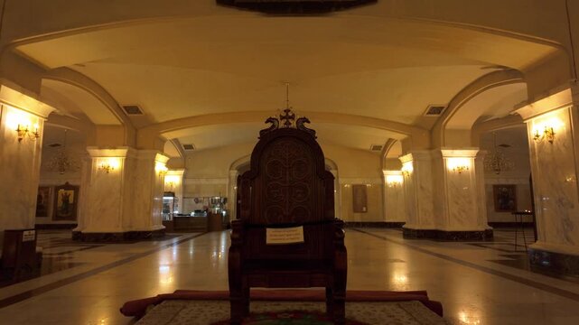 Ornate wooden throne centrally positioned within the vast hall of Holy Trinity Cathedral in Tbilisi. Marble pillars, archways, and ambient chandeliers define the sacred space.