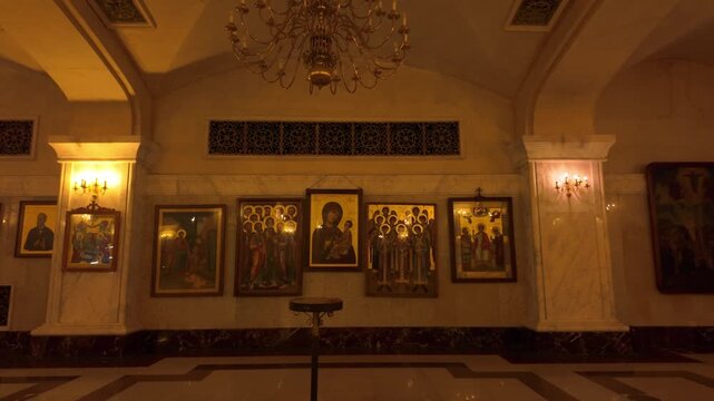 Beautifully illuminated wall section inside the Holy Trinity Cathedral in Tbilisi, featuring a row of significant Orthodox icons, a grand chandelier, and decorative architectural elements.