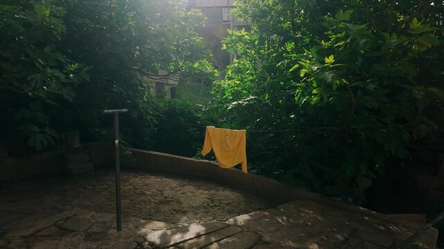 Bright yellow shirt hanging on a clothesline, drying in dappled sunlight amidst a lush, overgrown garden area. Cobblestone steps lead downwards, surrounded by dense green trees and vegetation creat...