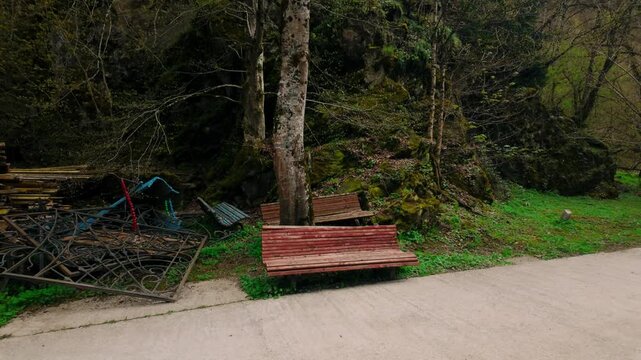 Concrete path with an empty red bench in a park setting. Beside it lies a pile of discarded materials including metal fences and wooden planks, backed by a lush, moss-covered rock face and dense tr...