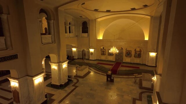 Panoramic high-angle view of the main hall inside Holy Trinity Cathedral, showcasing the altar area, numerous icons, and ornate chandeliers. Golden light illuminates the revered space and intricate...
