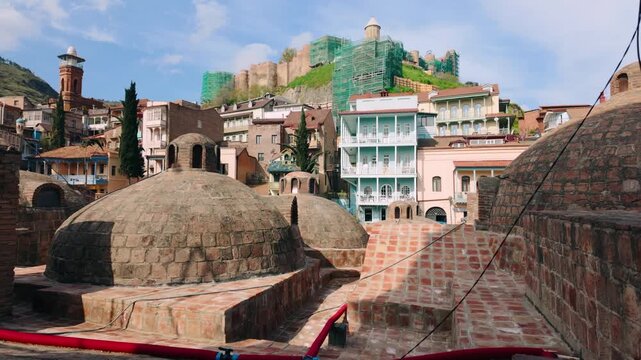 Iconic domed sulfur bathhouses of Abanotubani stand prominent against a backdrop of colorful traditional homes and the historic Narikala Fortress undergoing restoration in Tbilisi, Georgia.