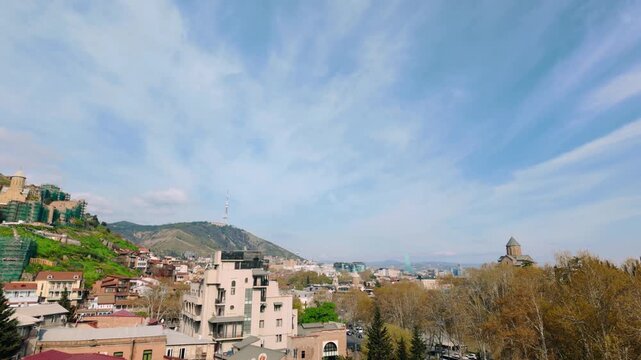 Wide daytime cityscape of Tbilisi, Georgia, offering a view of the Narikala Fortress and the TV tower, surrounded by a mix of historic and modern residential buildings and lush green areas.
