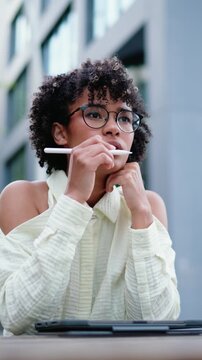 Focused dark-skinned digital nomad working remotely on touchpad with stylus in urban setting. Represents flexible, tech-driven lifestyle, embracing mobility and digital connectivity, freelancing in ci
