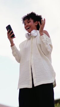 Excited Black woman receiving positive good news on smartphone, reacting joyfully while holding phone device. Celebrates notification with headphones around neck. Copy space, open sky background

