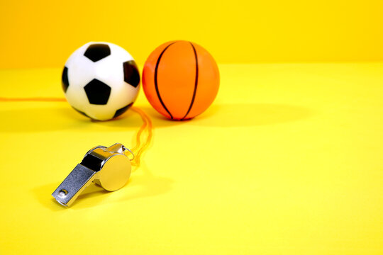 A metallic whistle with orange lanyard rests on a bright yellow surface next to a miniature soccer ball and a small basketball, evoking a sense of sports and competition. Yellow background.