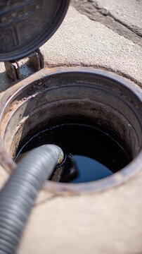 Close view of a manhole with a flexible hose entering dark liquid in a dirty concrete ground during daylight in urban settings