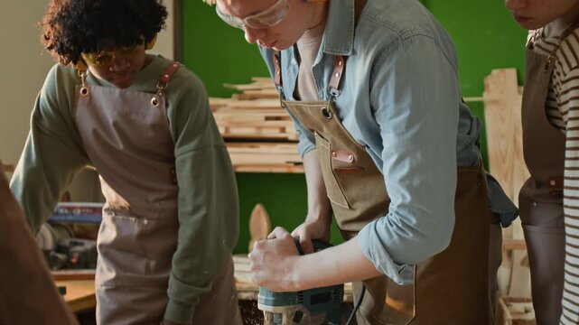 Tilt up shot of professional young carpenter using power saw while showing teenage students how to work with wood during masterclass