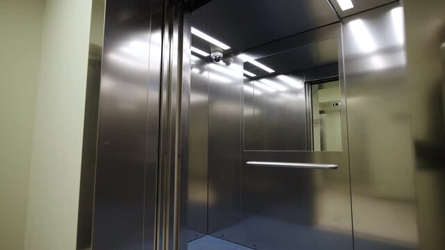Elevator interior includes polished steel walls and parallel ceiling lights. Integrated security dome camera ensures constant surveillance