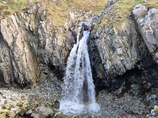 A small waterfall cascades through a narrow rocky gorge surrounded by grassy slopes and rugged stone formations in a natural outdoor landscape. © Studio-M