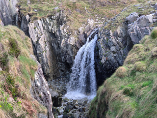 A small waterfall cascades through a narrow rocky gorge surrounded by grassy slopes and rugged stone formations in a natural outdoor landscape. © Studio-M