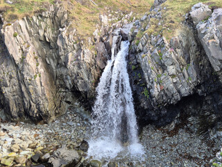 A small waterfall cascades through a narrow rocky gorge surrounded by grassy slopes and rugged stone formations in a natural outdoor landscape. © Studio-M