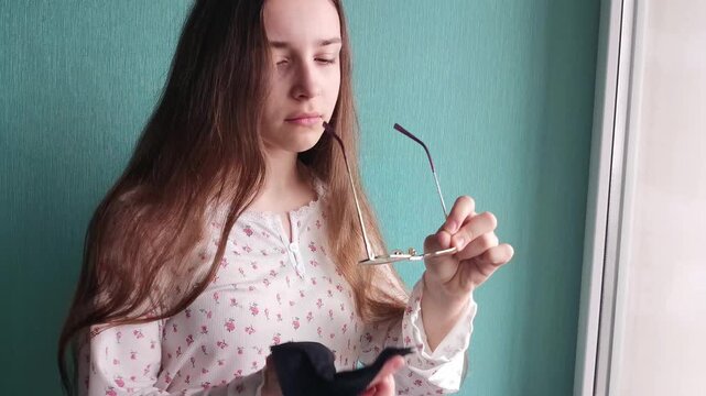 Teen girl cleaning eyeglasses with cloth indoors, vision care routine, lifestyle and personal maintenance concept.